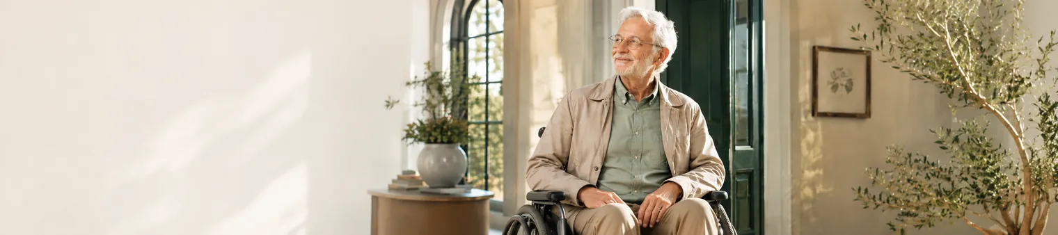 A patient using a wheelchair, calmly looking out a sunlit window in his home.